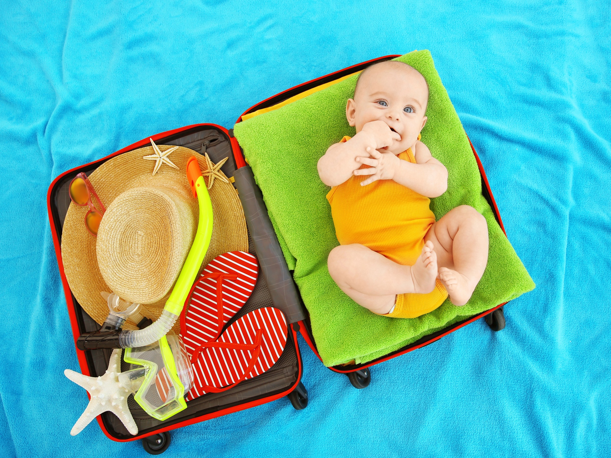 Cute baby and travel essentials in suitcase lying on blue bedspread